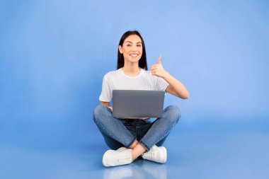 Eğitici kavram. Heyecanlı kız dizüstü bilgisayarla yerde oturuyor, baş parmak işareti yapıyor, mavi stüdyo duvarı.