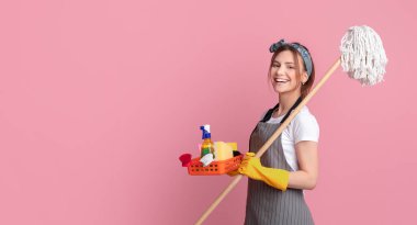 Profesyonel Temizlik. Gülümseyen genç kadın portresi, ev eşyaları ve el aletleriyle pembe arka planda poz vererek kameraya bakıyor.