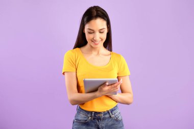 Teknoloji Konsepti. Mor stüdyo arka planında izole edilmiş dijital tablet kullanan mutlu genç bayan portresi. Gülümseyen kadın sosyal ağlara göz atıyor, haberleri okuyor, modern aletlerden hoşlanıyor.