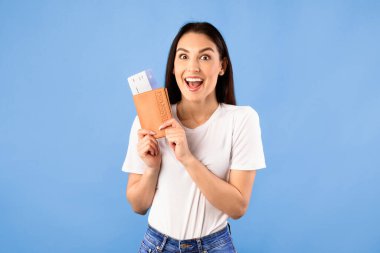 Ucuz Uçuş Konsepti. Bilet ve pasaport taşıyan, turkuaz stüdyo duvarında duran çok mutlu genç bir kadın.
