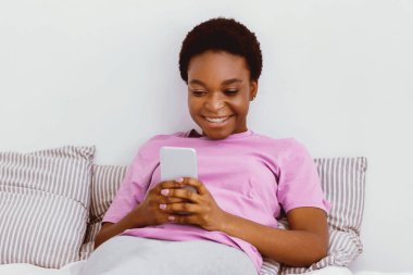 Evde internette sörf yapmak. Gülümseyen Afrikalı Amerikalı kız yatakta uzanıyor ve akıllı telefon kullanıyor.