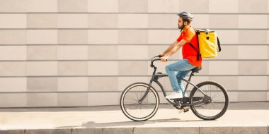 Gri duvar arka planında kurye bisikletçi. Sakallı, koruyucu kasklı, sarı sırt çantalı, müşteriye giden yolda, kenar manzaralı, serbest alan