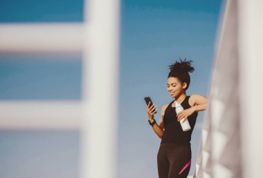 Sosyal medya ve spor. Gülümseyen Afro-Amerikan bayan spor kıyafetleri içinde elinde bir şişe suyla stadyumdaki akıllı telefona bakıyor.