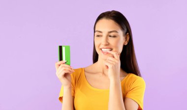 Hayalperest kız elinde banka kartına bakıp dudaklarının yanında parmağını tutuyor. Alışverişi düşünüyor. Mor renkte bir duvar.