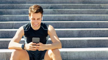 Rahat spor için modern cihazlar. Spor kıyafetli, kablosuz kulaklıklı ve spor takip cihazlı gülümseyen bir adam şehrin merdivenlerinde oturuyor ve akıllı telefonunda yazıyor.