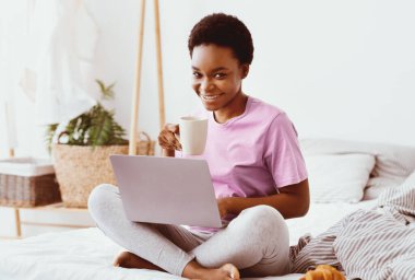 Evde uzaktan çalışma, blog, yatakta kahvaltıda sosyal medya. Mutlu binlerce yıllık Afro-Amerikan kadını dizüstü bilgisayarında bir fincan sıcak içecekle sabahları yatak odasının iç kısmında kruvasanla yazışıyor.