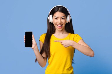 En iyi müzik uygulaması. Kulaklıklı neşeli Asyalı kız siyah ekranlı akıllı telefonu işaret ediyor Violet Stüdyo Arkaplanı 'nın üstünde duruyor, Mockup Image