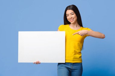 Reklamınız için yer. Gülümseyen Asyalı kız elinde beyaz boş levha, fotokopi uzayını işaret eden, Mor Stüdyo Arkaplanı Üzerine Poz Veren