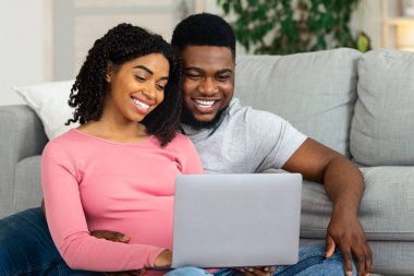 Hamile Afro-Amerikan çifti evde dizüstü bilgisayar kullanarak, oturma odasında oturup, bebek odası iç mekanlarını izleyerek sevmek. Gülümseyen siyah adam ve kadının dizüstü bilgisayara bakmasını beklemek.