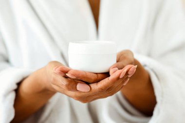 Bornozlu, tanınmayan siyah bir kadının ellerinde bir kavanoz besleyici krem, evde cilt bakımı, yakın çekim...