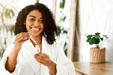 Bornozlu mutlu afro kadın elinde organik serumla duştan sonra güzel yüzüne sürmeye hazır.