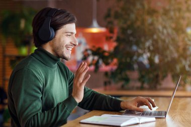 Kablosuz kulaklıklı mutlu adam kafede online konferans veriyor, dizüstü bilgisayar kullanıyor, ekrana el sallıyor, boşluğu kopyalıyor, yan görünümü kopyalıyor.