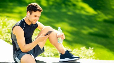 Eğitimden sonra dinlenin. Kablosuz kulaklıklı gülümseyen adam akıllı telefona bakıyor, içinde su olan şişeyi tutuyor, parkta oturuyor, boş yer var.