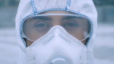 Genç bir Orta Doğulu kadın dışarıda duruyor koruyucu giysiler ve maske takıyor. Odaklanmış bakışları ciddiyetini gösteriyor, mevcut halk sağlığı endişelerinin özünü yakalıyor..