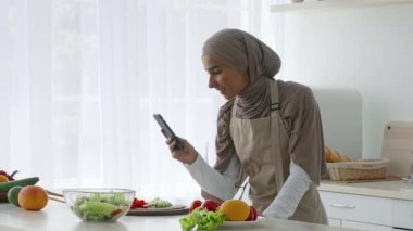 Orta Doğulu tesettürlü bir kadın açık bir mutfakta yemek hazırlamakla meşgul. Etrafı taze sebze ve meyvelerle çevriliyken telefonunu kontrol ediyor..