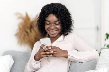 Yurtiçi Dinlenme. Akıllı telefonlu rahat koltukta rahatlayan, internete göz atan, arkadaşlarıyla mesajlaşan, cep telefonuyla vakit geçiren ve hafta sonunun keyfini çıkaran mutlu siyah kadın.