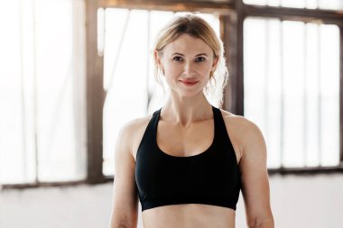 Kaslı, güçlü ve güzel bir vücut. Spor kıyafetleri içinde gülümseyen orta yaşlı bir kadın. Pencere arkasında spor saati ve eldivenli. Kopya alanı.