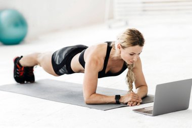 Coronavirus salgını sırasında evde online ders. Spor üniformalı çekici, olgun bir sporcu yerde spor topu ve su şişesiyle döşeme tahtası yapıyor ve dizüstü bilgisayara bakıyor.