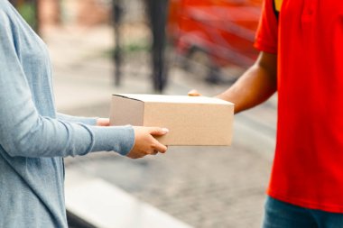 Eve paket teslimatı, sipariş ve kurye servisi. Turuncu üniformalı akıllı bir adam sokakta, evin yanında, manzaralı, panoramada, boş alanda kutusunu kadına veriyor.