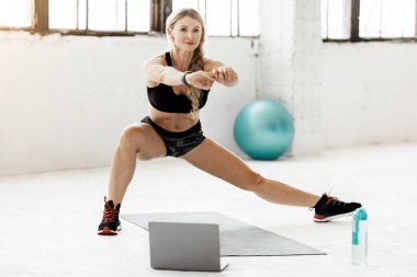 Covid-19 salgını ve kendini izole etme sırasında evde online egzersiz. Spor kıyafetleri giymiş sakin yetişkin bir kadın dizüstü bilgisayarla tavan arasında bir şişe su ve fitness topu ve bedava uzay topuyla hamlesini yapıyor.