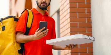 Şehirde kuryeyle pizza teslimatı. Kasklı ve sarı çantalı ciddi bir adam, içinde yiyecek ve saatlerin olduğu karton bir kutu tutuyor. Akıllı telefon, ön kapı, kopyalama alanı.