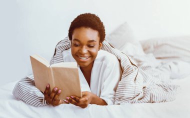 Sabah kitap okuma. Mutlu Afrikalı Amerikalı kadın battaniyeye sarılmış, güneş parlıyor