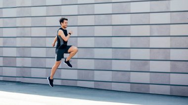 Zıplayarak koşuyor. Adam gün ışığında gri tuğla duvarın yanındaki yolda koşuyor.