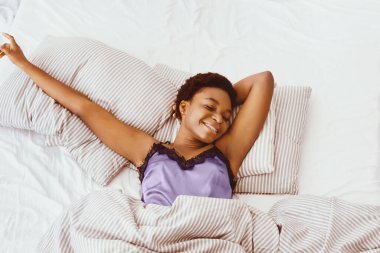 Tembellik ve günaydın, Pazar sabahı uyu ve tatilin tadını çıkar. Mutlu Afrikalı genç Amerikalı bayan yatağa uzanıyor ve yumuşak yastıklara uzanıyor, battaniyeli beyaz çarşaf, üst manzara, fotokopi odası