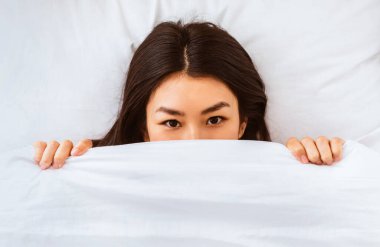 Oyuncu Asyalı kadın battaniyeden dışarı bakıp sabah yatağında yatarak uyanıyor. Üst Görünüm