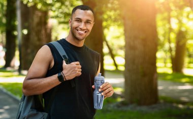 Parktaki eğitiminden sonra sırt çantası ve su şişesiyle gülümseyen siyah sporcunun portresi.