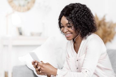Evde boş zaman. Smartphone 'da müzik dinleyen, kulaklık takan, video izleyen, oturma odasında dinlenirken interneti kolaçan eden, hafta sonunun keyfini çıkaran mutlu siyah kadın.