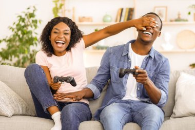 Video oyunu oynayan mutlu siyah çift, erkek arkadaşının gözlerini kapatan kadın, ev içi.