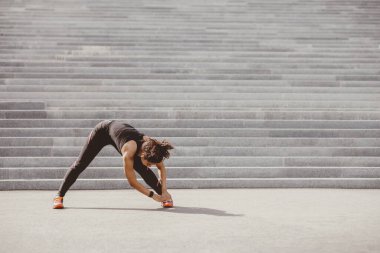 Egzersiz sonrası esneme hareketleri. Spor kıyafetli, spor ayakkabılı, kablosuz kulaklıklı, eğik bacaklı şehir merdivenleri yakınında egzersiz yapan Afrikalı Amerikalı bir kadın.