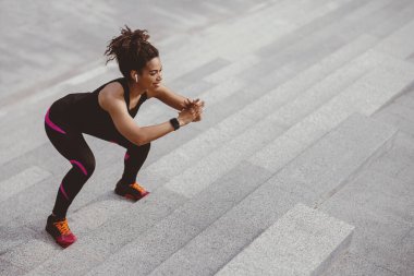 Zıplıyor ve merdivenlerde çömeliyor. Kablosuz kulaklıkları ve spor aleti olan gülümseyen Afrikalı Amerikalı kız şehirde merdivenlerde egzersiz yapıyor.