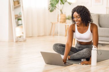 Neşeli Afrikalı kadın evde egzersiz yapıyor, dizüstü bilgisayar kullanıyor, fitness videosunu seçiyor, boş alan seçiyor.