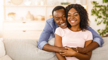 Neşeli Afrikalı bir adam, gülümseyen karısına arkadan sarılıyor, evde birlikte vakit geçiriyor, boş bir alanda.