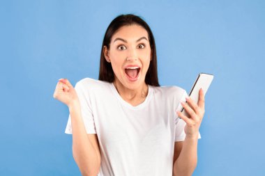 Öyle mi? Mavi stüdyo arka planında izole edilmiş, cep telefonu kullanan mutlu genç bir kadının portresi. Heyecanlı bayan online zaferi kutluyor, çığlık atıyor ve kameraya bakıyor.