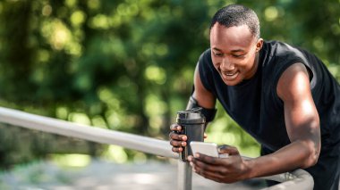 Gülümseyen Afro-Amerikan sporcu telefonda konuşuyor, protein içiyor, parkta egzersiz yapıyor, boş alan var.