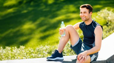 Sağlık ve spor. Spor kıyafetli gülümseyen adam elinde bir şişe suyla parkta oturuyor.