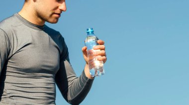 Spor için su. Spor kıyafetlerinde yorgun adam elinde şişeyle mavi gökyüzü arka planında, boş alanda koşuyor.