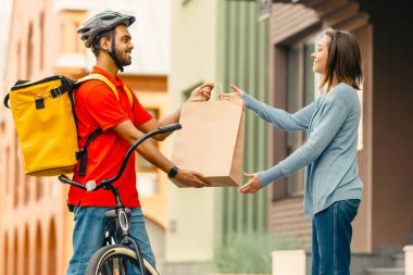 Yemek, paket teslimat konsepti. Gülümseyen milenyum sakallı Hintli erkek, güvenlik kasklı, üniformalı, sarı termal çantalı akıllı saat ev, yan görüş, panorama yakınlarındaki kadın müşteriye paket veriyor.