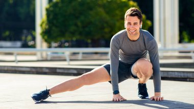 Parkta koşmadan önce ısın. Spor üniformalı genç adam bacak egzersizi yapıyor, açık havada, serbest alanda.