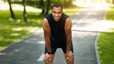 Güneşli parkta maraton koşusundan sonra dinlenen gülümseyen siyah koşucu,