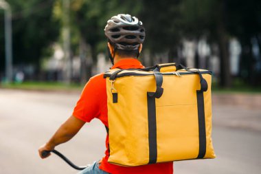 Kasabada kurye var. Koruyucu miğferli teslimatçı, büyük çantalı. Yolda bisiklet sürüyor. Şehirde, arkada, kopyalama alanında.