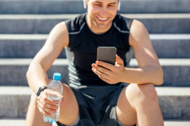 Yorgunluk ve antrenmanın sonu. Kablosuz kulaklıklı genç adam merdivenlerde oturuyor, elinde bir şişe su tutuyor ve akıllı telefona bakıyor.