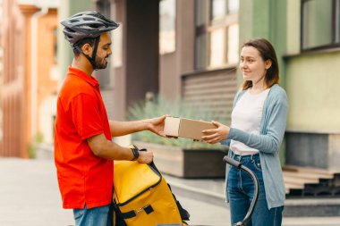 Memnun müşteri çevrimiçi sipariş alıyor. Kız, sarı sırt çantalı ve bisikletli kuryeden karton paketi alıp sokağa, yan manzaraya, boş alana götürüyor.