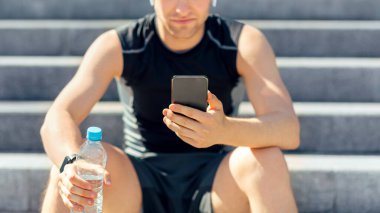 Cihazlar ve fiziksel aktivite. Spor kıyafetli, kulaklıklı yorgun adam akıllı telefonlara bakıyor ve su tutuyor, merdivenlerde oturuyor, boş yer var.