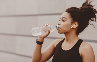 Çapraz, kardiyo eğitimi ve sağlıklı yaşam tarzı. Modern kulaklıklı ve fitness takipçisi olan kaslı Afrikalı Amerikalı kız gri duvar arka planında su içiyor, profil, kapanış, boşluk kopyalama