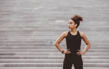 Eğitimin başındaki bir atlet portresi. Kablosuz kulaklıkları ve akıllı saati olan, ciddi bir Afrikalı Amerikalı kız şehrin arka planında duruyor ve boş bir yere bakıyor.