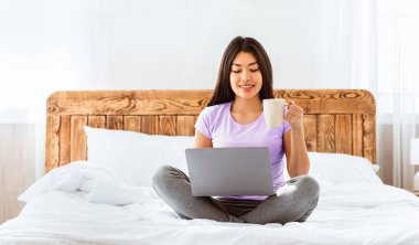 Asyalı kız dizüstü bilgisayar kullanıyor elinde kahve fincanı, hafta sonu evde yatağında oturuyor. Boş Alan